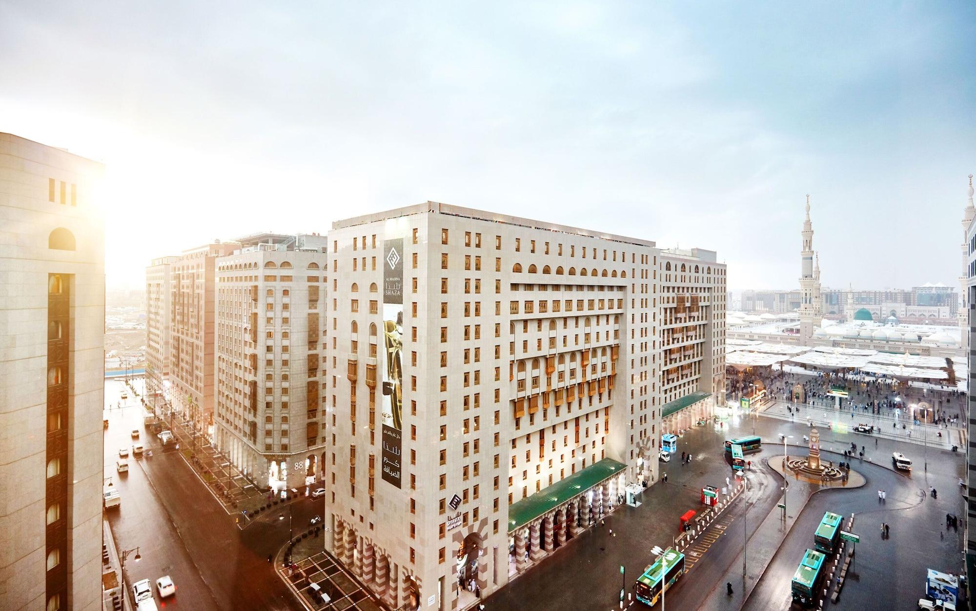 샤자 마디나 호텔 메디나 외부 사진 The photo shows an urban landscape featuring several large buildings, which appear to have a modern architectural style. The scene is depicted during a time of day when sunlight is visible, suggesting either early morning or late afternoon. In the fo