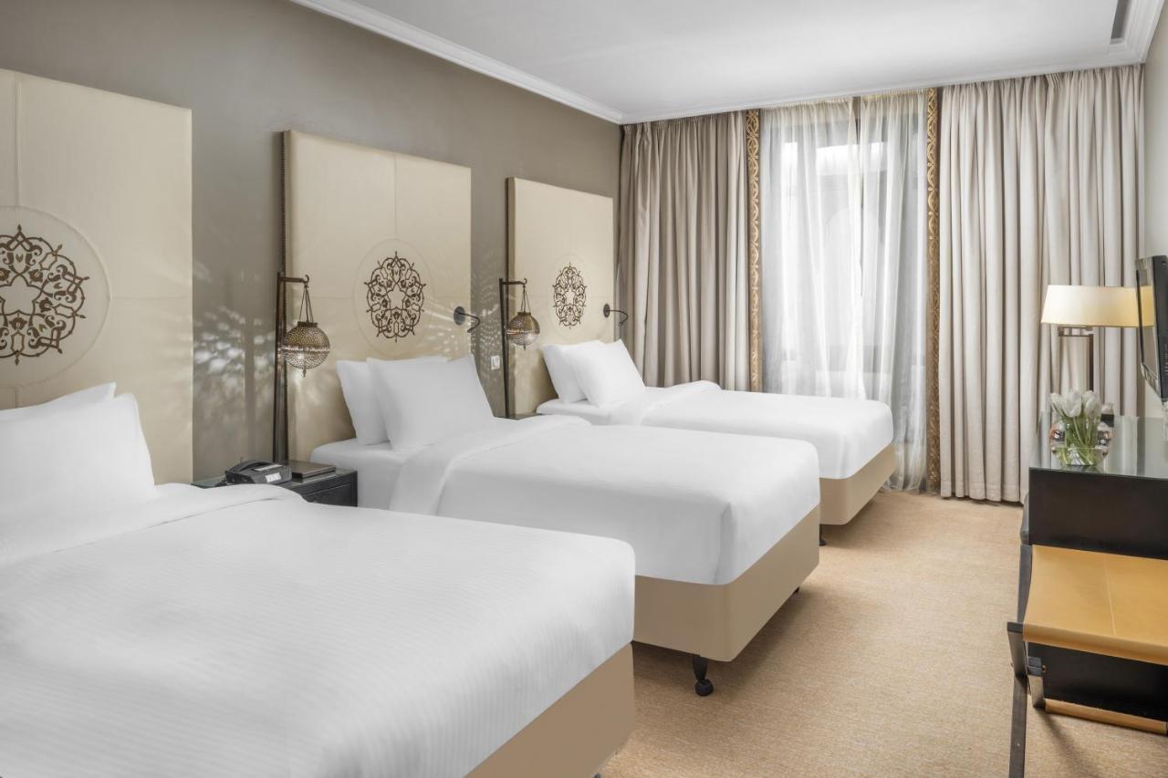샤자 마디나 호텔 메디나 외부 사진 The photo shows a hotel room featuring three beds, each made with white bedding. The beds are positioned against a light-colored wall adorned with decorative headboards. There are two bedside tables with lamps next to the beds. The room is well-lit, 