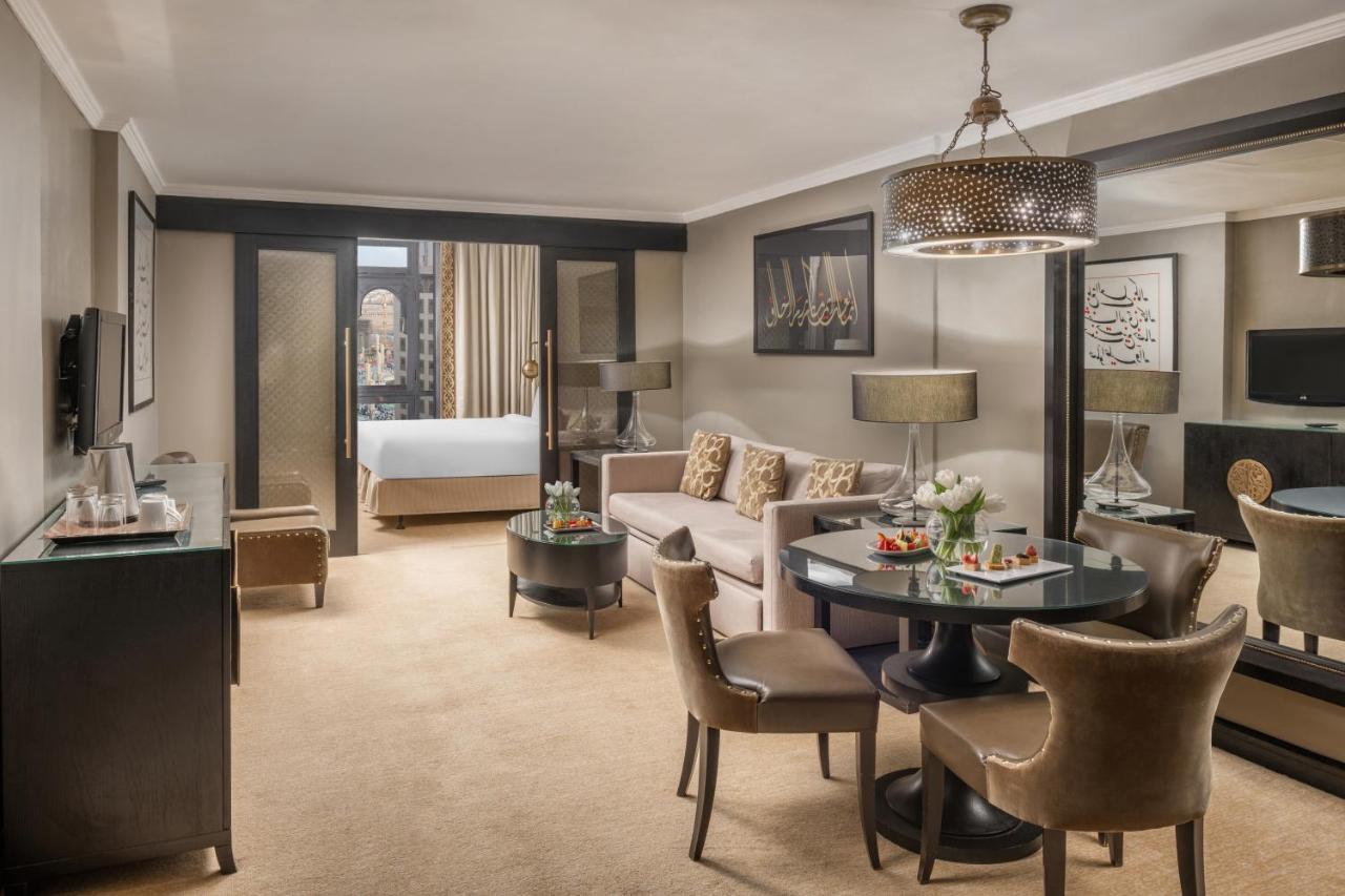 샤자 마디나 호텔 메디나 외부 사진 The photo shows a well-appointed hotel suite. It features a cozy living area with a beige sofa and several decorative cushions. A circular table with chairs is set in the center, likely for dining or working. There is a decorative chandelier overhead