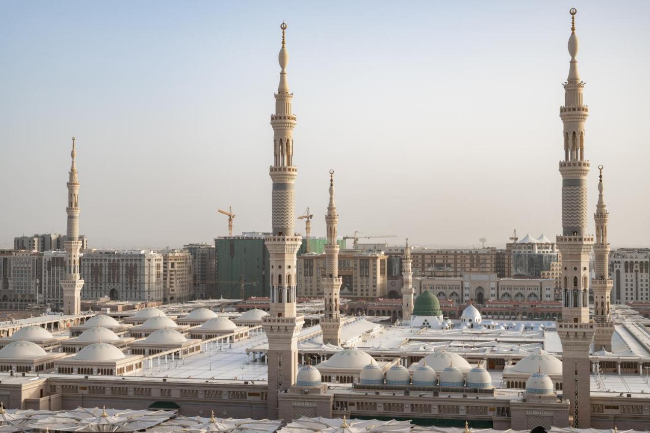 샤자 마디나 호텔 메디나 외부 사진 The photo depicts a view of an Islamic mosque complex with several prominent minarets. The architectural style features a blend of intricate designs and large, domed roofs. The scene appears to be set in a modern urban environment, with construction 
