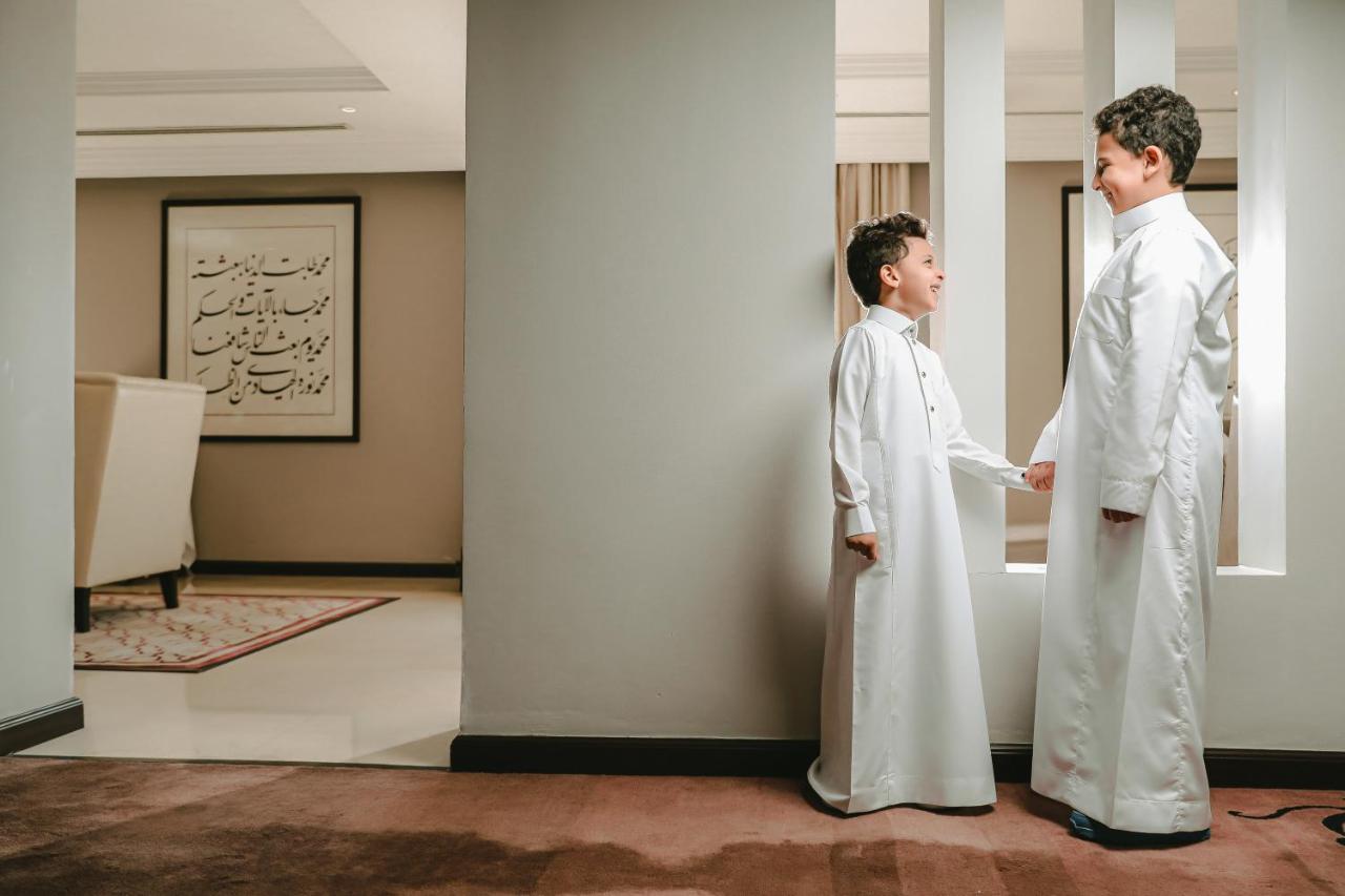 샤자 마디나 호텔 메디나 외부 사진 The photo shows two young boys dressed in traditional white clothing, likely thobes. They are standing in a hallway, engaged in a conversation or interaction. The setting appears to be an indoor space with modern decor, including a piece of artwork o