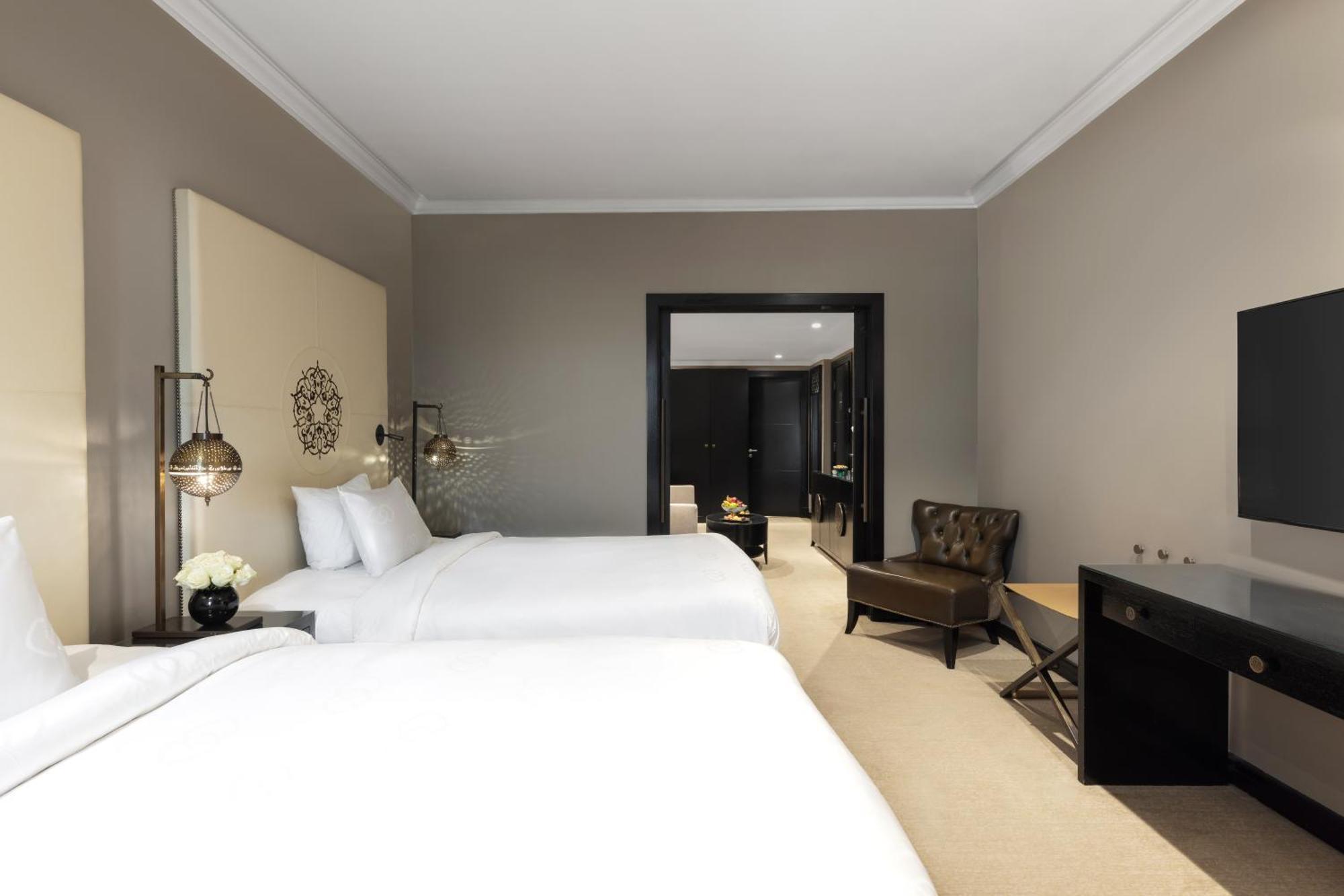 샤자 마디나 호텔 메디나 외부 사진 The photo shows a hotel room with a modern design. It features two white beds with neatly arranged pillows, a simple beige wall with decorative accents, and stylish bedside lamps. There is a small seating area with a brown chair and a side table, as 