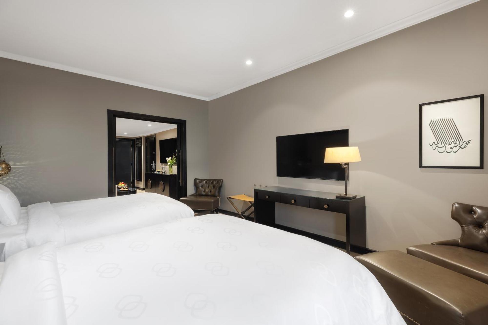 샤자 마디나 호텔 메디나 외부 사진 The photo shows a modern hotel room with a neutral color palette. It features two beds with white linens, a stylish lamp on a side table, and a flat-screen TV mounted on the wall. There is a dark wood console table beneath the TV, and a comfortable a