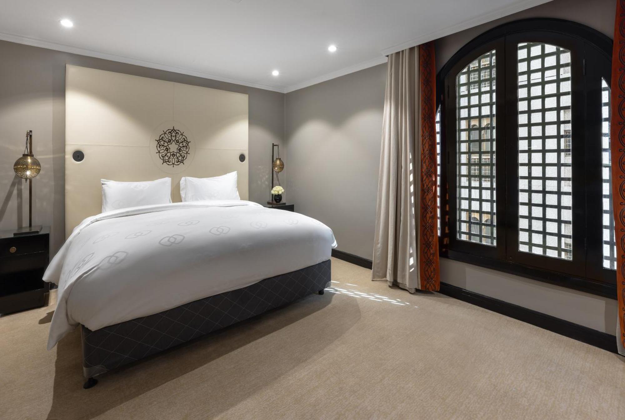 샤자 마디나 호텔 메디나 외부 사진 The photo shows a modern hotel bedroom. It features a large bed with white bedding and two pillows. The walls are painted in neutral tones, and there is decorative wall art above the bed. To the right, there are large windows with patterned shutters,