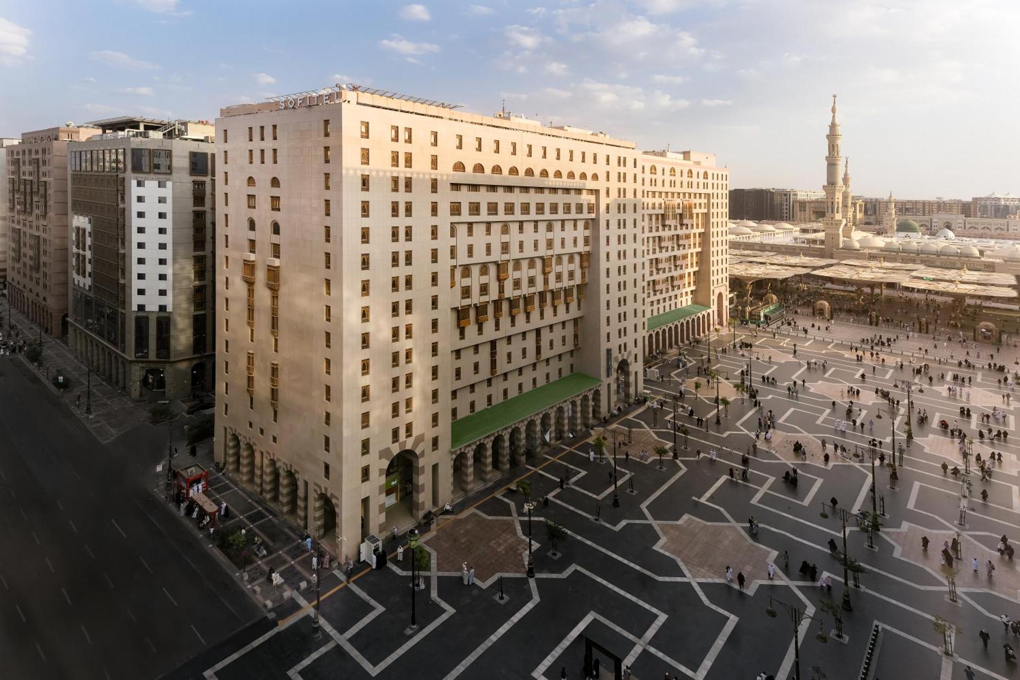 샤자 마디나 호텔 메디나 외부 사진 The photo shows a large, multi-story building with a distinct architectural design, located near a spacious outdoor plaza. The building features a combination of columns and large windows, giving it a formal appearance. In front of the building, ther