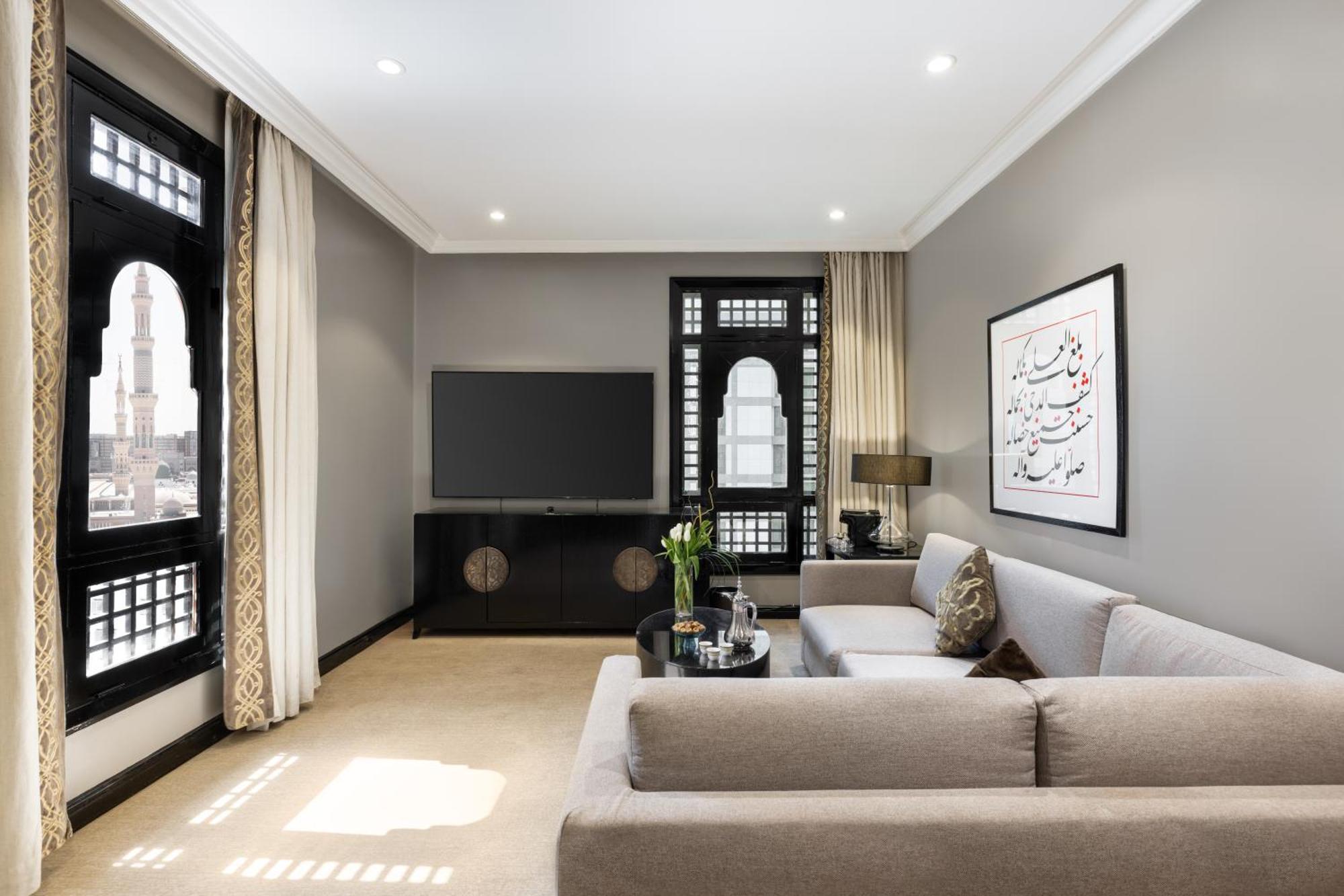 샤자 마디나 호텔 메디나 외부 사진 The photo depicts a modern living room with a neutral color palette. There's a comfortable light gray sofa with decorative cushions positioned toward the center. Opposite the sofa, a sleek black TV unit supports a flat-screen television. Large window