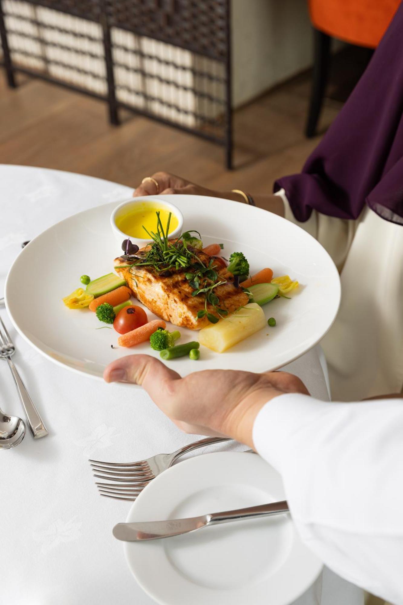 샤자 마디나 호텔 메디나 외부 사진 The photo shows a plate of food being presented. The dish features a piece of grilled salmon on a bed of vegetables, including small tomatoes, green peas, and other colorful vegetables. There is also a drizzle of yellow sauce, likely a dressing or oi